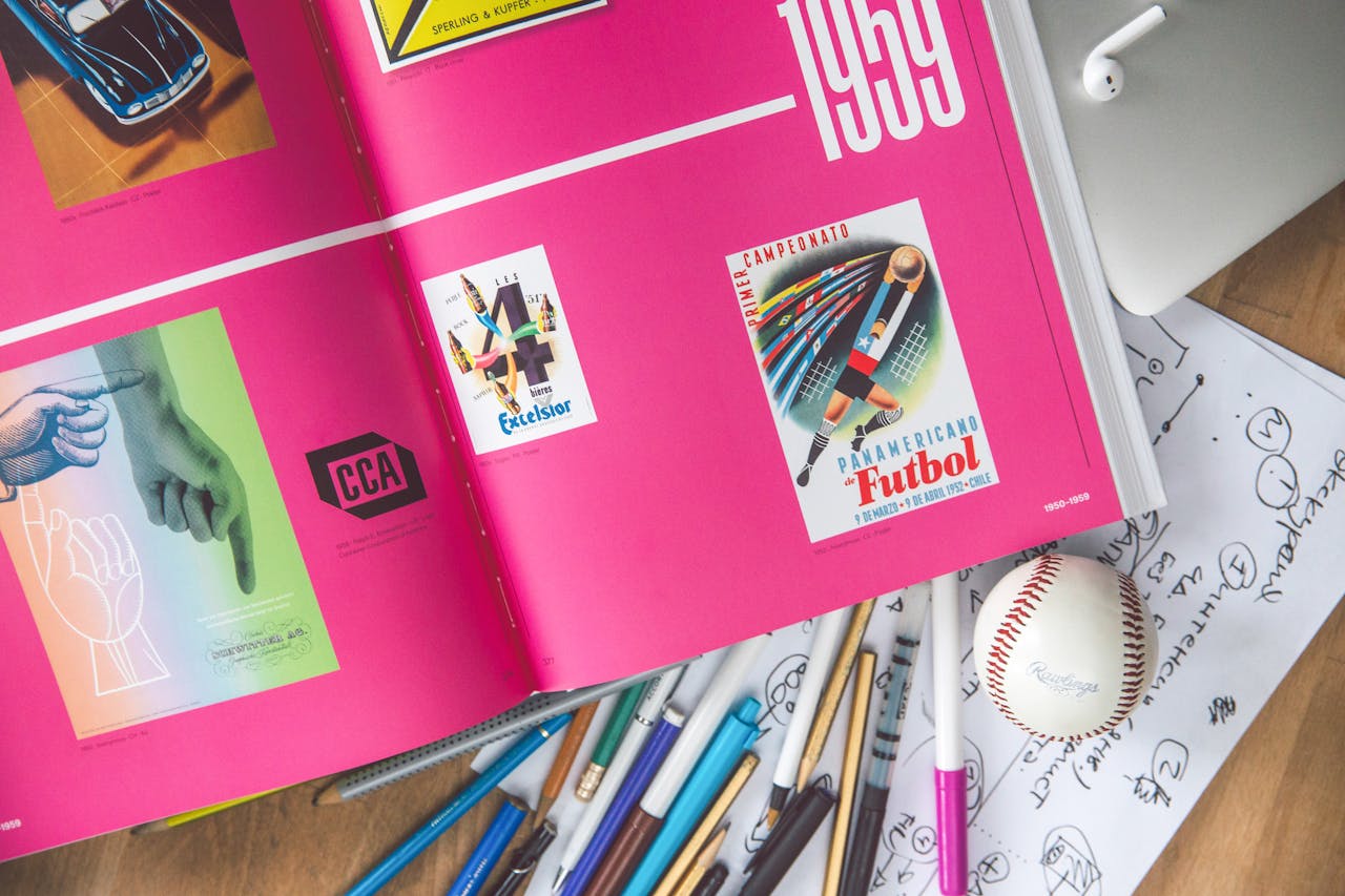 Artistic workspace featuring an open design book, sketches, pens, and a baseball on a desk.